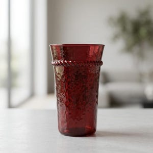 May include: A deep red glass tumbler featuring a textured, hammered surface and a decorative rope-style band encircling the upper rim. The vessel has a slightly flared opening and a cylindrical body, crafted from translucent, colored glass material.