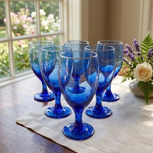 May include: Six cobalt blue wine glasses with a classic stem design are arranged on a table. The glasses are empty and sit on a white linen runner. A window and a floral arrangement are in the background.