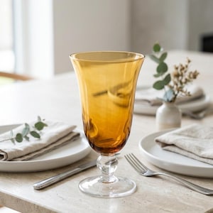 May include: A tall, amber-colored glass goblet with a smooth, flared bowl and a clear, round pedestal base. The stem features a small, spherical decorative accent just below the bowl, connecting to the transparent, circular foot of the glass.