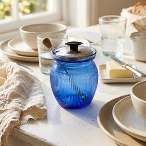 May include: A vibrant blue glass jar with a lid and a small spoon inside. The jar has vertical ribbed detailing and sits on a white table, alongside other tableware and a glass of water. The scene is bathed in natural light.