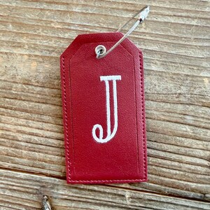 May include: A red leather luggage tag with a white embroidered letter "J". The tag is rectangular with a rounded top and is attached to a metal loop. The tag is on a wooden surface.