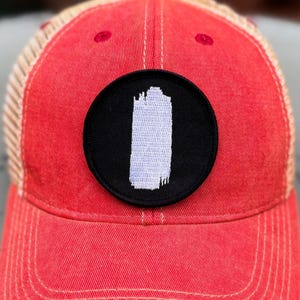 May include: A red baseball cap with a black circular patch featuring a white, vertical design. The cap has a mesh back and a curved brim. The text "(Hat Not Included)" is in the upper right corner.