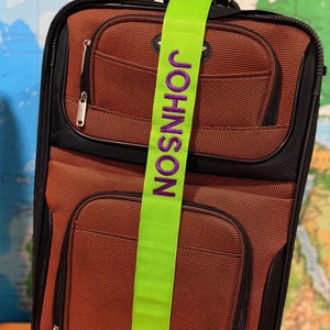 May include: A lime green luggage strap with the name "JOHNSON" embroidered in purple lettering. The strap is attached to a brown and black suitcase.