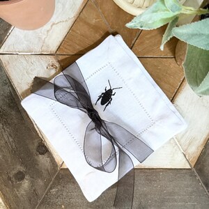 May include: White square linen cocktail napkin with a black embroidered insect design. The napkin is tied with a sheer black ribbon. The napkin is a decorative item.