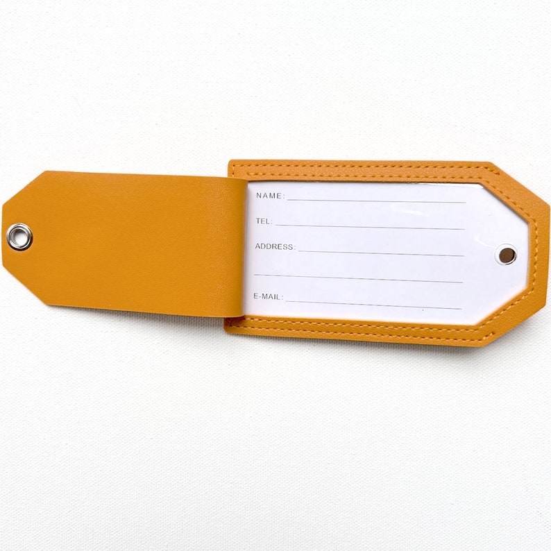 May include: A mustard yellow leather luggage tag with a white label that has the words "NAME", "TEL", "ADDRESS", and "E-MAIL" printed on it. The tag has a flap that folds over the label to protect the information.