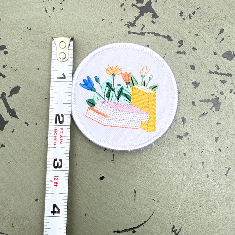 May include: Embroidered patch with a white border featuring a floral design with blue, orange, and white flowers, a pink book, and a yellow book. The patch is approximately 2 inches in diameter.