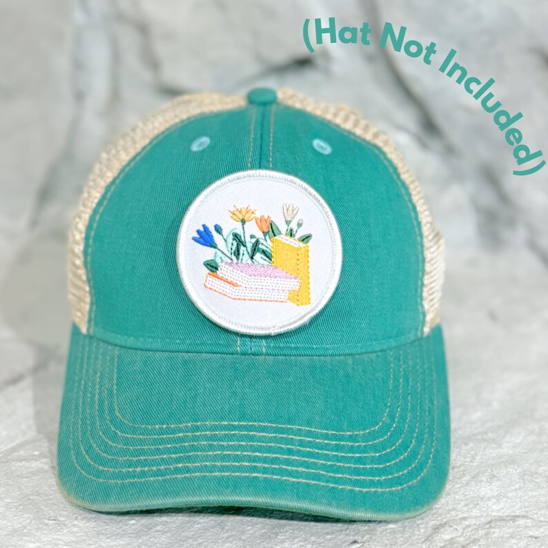 May include: Teal trucker hat with a beige mesh back and a circular patch. The patch features an embroidered design of books and flowers in yellow, blue, and orange. The text "(Hat Not Included)" is in the upper right corner.
