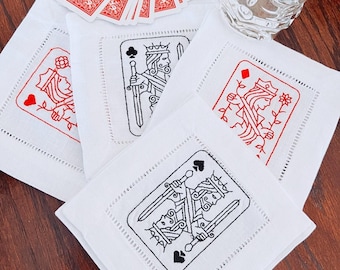 May include: Four square white fabric coasters featuring embroidered playing card designs. Each coaster displays a different face card, including the King of Clubs, Queen of Hearts, Queen of Diamonds, and King of Spades, with hemstitched borders.