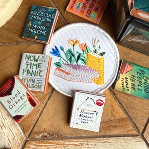 May include: A collection of miniature books and an embroidered patch with a floral design. The books have titles like "The Minuscule Mansion" and "Now Is Not the Time to Panic." The patch features colorful embroidered flowers.