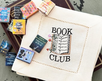 May include: A square cream-coloured fabric napkin featuring an embroidered open book with flowers and the text "BOOK CLUB" in black thread. Surrounding the napkin are several miniature book replicas with titles including "The Authenticity Project," "The Measure," "Pretty Girls," and "The Push."