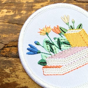 May include: Embroidered patch featuring a bouquet of flowers in shades of orange, blue, and yellow, with green leaves and a yellow container. The patch has a white border and is on a wooden surface.