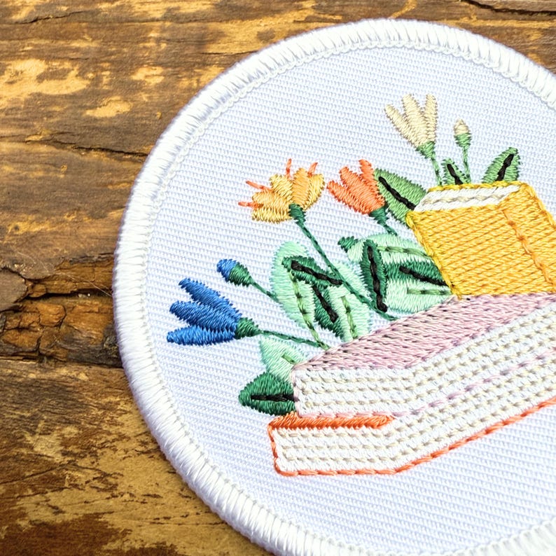 May include: Embroidered patch featuring a bouquet of flowers in shades of orange, blue, and yellow, with green leaves and a yellow container. The patch has a white border and is on a wooden surface.