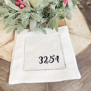 May include: A square white linen napkin featuring a decorative hemstitch border and the black embroidered number 3254 centered on the fabric. The napkin is positioned beneath a festive wreath with green foliage and red berries.