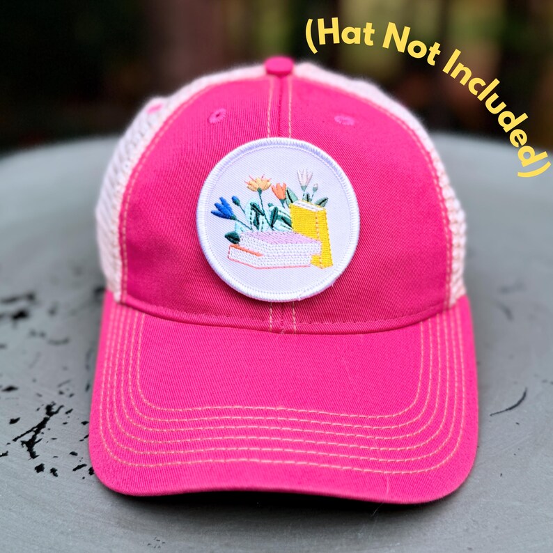 May include: A bright pink trucker hat with a white mesh back and a circular embroidered patch. The patch features a stack of books and flowers in shades of blue, orange, and yellow. The text "(Hat Not Included)" is in the upper right corner.