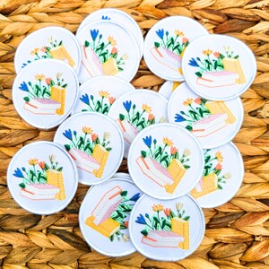 May include: A collection of white, circular embroidered patches. Each patch features a stack of books with colorful flowers and foliage. The books are yellow and pink, and the flowers are blue, orange, and yellow. The patches are arranged on a woven, brown surface.