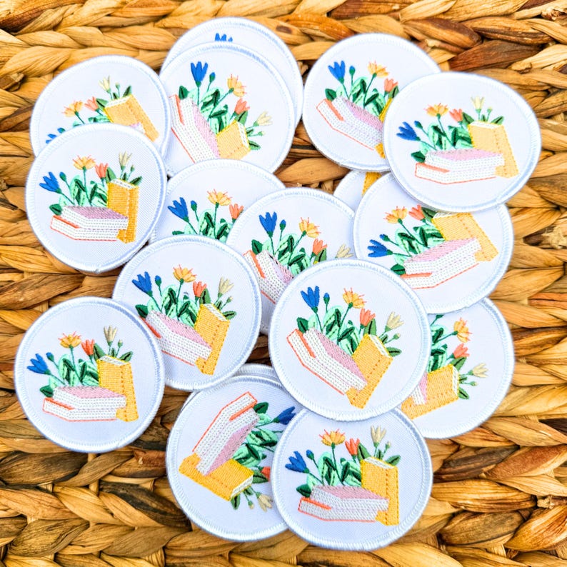 May include: A collection of white, circular embroidered patches. Each patch features a stack of books with colorful flowers and foliage. The books are yellow and pink, and the flowers are blue, orange, and yellow. The patches are arranged on a woven, brown surface.