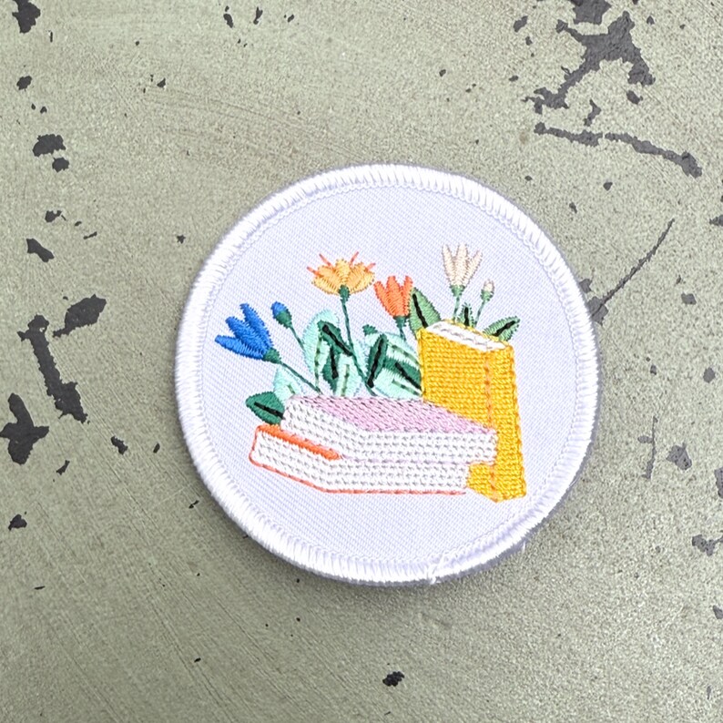 May include: Embroidered patch with a white border featuring a floral and book design. The design includes blue, orange, and white flowers, with green leaves and two books in the center. The books are yellow and pink.