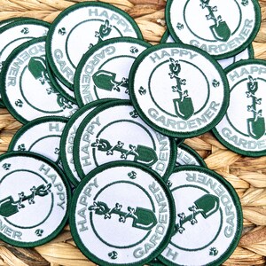 May include: A collection of round white patches with a green border. Each patch features the words "HAPPY GARDENER" in a circular design around a green shovel and plant illustration. The patches are scattered on a woven surface.
