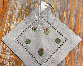 May include: A square light gray linen cocktail napkin featuring an embroidered border of openwork stitching and five small green olive motifs with red pimentos. A clear glass stemware base rests on the center of the fabric napkin.