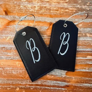 May include: Two black luggage tags with the letter "B" in a cursive font. Each tag has a silver-colored wire loop for attachment. The tags are displayed on a wooden surface.