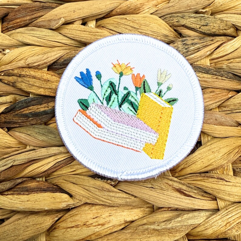 May include: Embroidered patch featuring a floral and book design. The patch is round with a white background and a stitched border. The design includes blue, orange, yellow, and white flowers, and two books in pink and yellow.