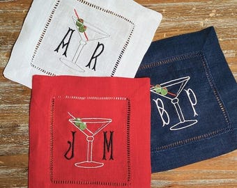May include: Three square linen cocktail napkins in white, red, and navy blue. Each napkin features an embroidered martini glass with green olives and two black letters. The white napkin shows A R, the red napkin shows J M, and the navy napkin shows B P.