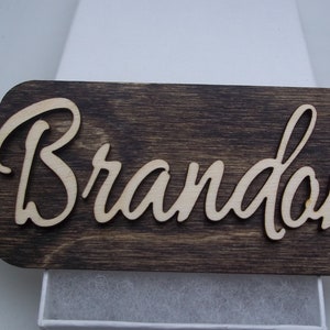 May include: A wooden name tag with the name "Brandon" cut out in a cursive font. The tag is dark brown with a distressed finish and has a hole at the top for hanging.