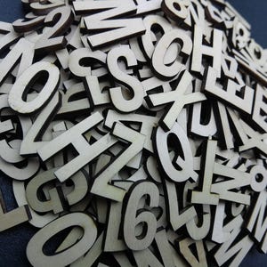May include: A large collection of individual, flat, light-colored wooden letters and numbers scattered in a dense pile. Each piece features a clean, sans-serif typeface cut from thin wood, showing natural grain textures and dark, laser-cut edges.