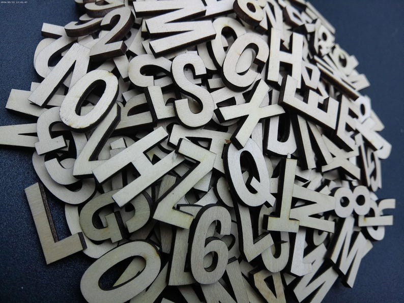 May include: A large collection of individual, flat, light-colored wooden letters and numbers scattered in a dense pile. Each piece features a clean, sans-serif typeface cut from thin wood, showing natural grain textures and dark, laser-cut edges.