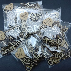 May include: Multiple clear plastic bags filled with small, light-colored wooden alphabet letters. Each bag is sealed with a heart-shaped tan paper sticker featuring the text "thank you" printed in a dark, cursive script font.