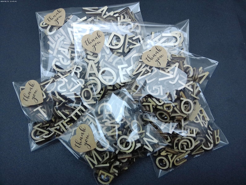 May include: Multiple clear plastic bags filled with small, light-colored wooden alphabet letters. Each bag is sealed with a heart-shaped tan paper sticker featuring the text "thank you" printed in a dark, cursive script font.