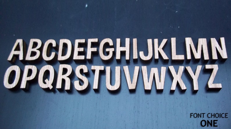 May include: A complete set of twenty-six individual wooden capital letters from A to Z arranged in two rows. Each letter features a light brown wood grain texture and a sans-serif typeface. The text FONT CHOICE ONE is printed in the bottom right corner.
