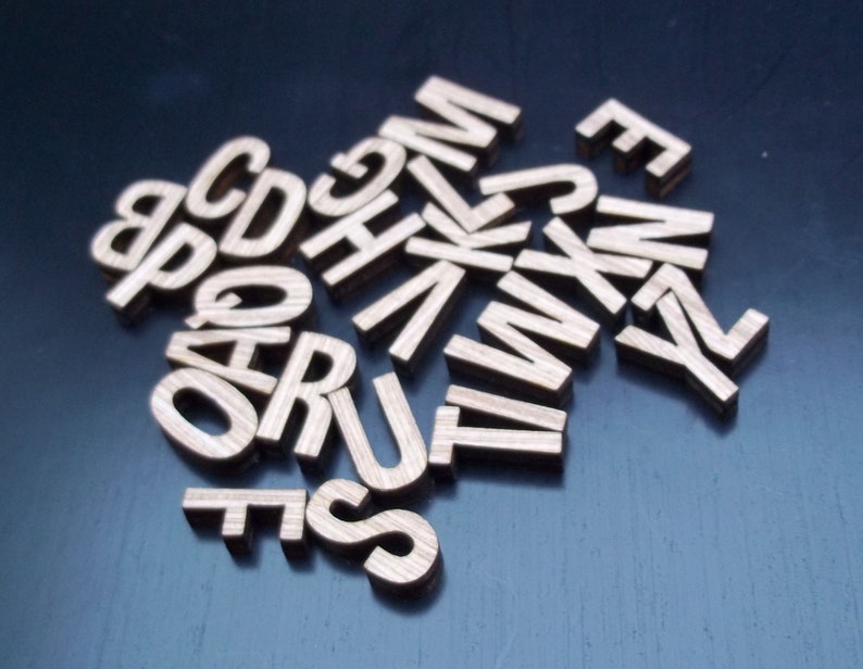 May include: A collection of individual wooden letters scattered in a pile. Each letter features a natural brown wood grain texture and a clean, sans-serif typeface. The letters are arranged in a random orientation, showcasing various characters of the alphabet.