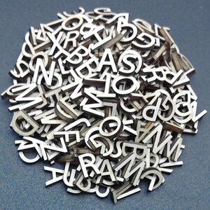 May include: A large collection of small, individual wooden letters of the alphabet. Each piece features a white front surface with dark, natural wood edges, creating a high-contrast appearance. The letters are scattered in a dense, overlapping pile.