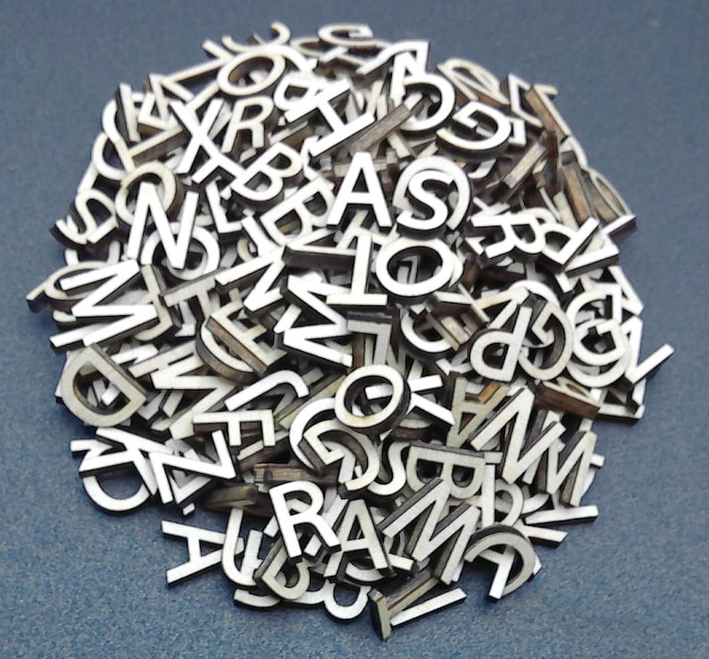 May include: A large collection of small, individual wooden letters of the alphabet. Each piece features a white front surface with dark, natural wood edges, creating a high-contrast appearance. The letters are scattered in a dense, overlapping pile.