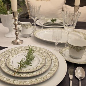 May include: Elegant white and green floral patterned china dinnerware set. Includes plates, a teacup and saucer, a creamer, and wine glasses. Silverware and a sprig of rosemary are also present. Two lit candles in silver candlesticks complete the table setting.