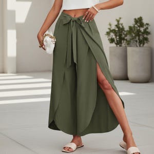 Op de afbeelding: Olijfgroene palazzo broek met wikkelstijl, hoge taille en striksluiting. De broek heeft een splitbeenontwerp en wordt gecombineerd met witte sandalen. Een witte clutch met een gouden ketting is ook zichtbaar.