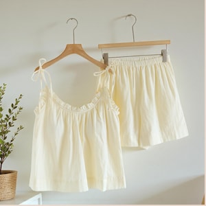May include: A cream-colored pajama set, consisting of a camisole top and shorts, hangs on wooden hangers. The top features tie straps and a ruffled neckline. The shorts have an elastic waistband. The set is made of a soft, lightweight fabric.