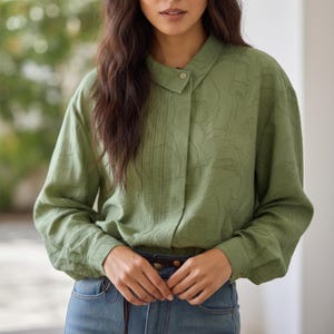 May include: A long-sleeved, olive green blouse with a mandarin collar and button closure. The fabric has a subtle, embossed floral pattern. The blouse features vertical pleats down the front and is paired with blue jeans.