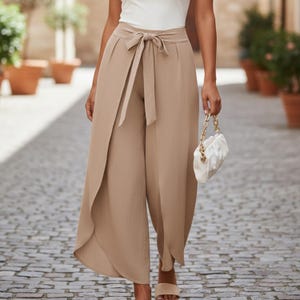 May include: Tan-colored wide-leg pants with a wrap-style front and a matching tie belt. The pants are paired with a white top and sandals. A white purse with a gold chain is held in the hand.