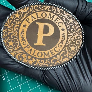 May include: A round, brown and gold belt buckle with the name "PALOMEC" and a large letter "P" in the center. The buckle has a decorative floral and star design, with a silver rope-like border.