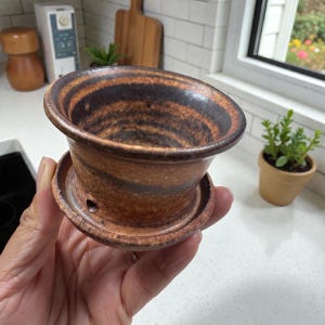 Puede incluir: Una maceta de cerámica marrón y negra hecha a mano con base de platillo. La maceta tiene un borde ancho y acampanado y un patrón arremolinado texturizado. Se puede ver un pequeño orificio de drenaje en el platillo. La maceta está diseñada para plantas pequeñas.
