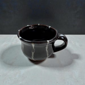 May include: A handmade, dark brown ceramic mug with a rounded body and a small handle. The mug features vertical white streaks and white speckles around the rim. The interior is a dark brown color. The mug is sitting on a light gray surface.