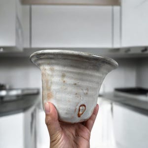 May include: A handmade ceramic bowl with a flared rim. The bowl is primarily white with gray and brown accents, and a unique, organic shape. The bowl is held in a hand, showcasing its size and texture. The bowl is likely for kitchen or home decor.
