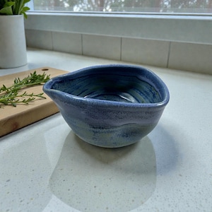 Op de afbeelding: Een handgemaakte, blauwe keramische kom met een unieke, asymmetrische vorm. De kom heeft een getextureerd oppervlak en een verloop van blauwe en groene tinten. Het wordt waarschijnlijk gebruikt voor het serveren van sauzen of kleine porties.