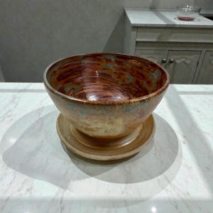 May include: A handcrafted ceramic bowl with a wide, rounded shape, resting on a matching base. The bowl features a rich, earthy color palette of brown, rust, and cream, with subtle blue-green accents. The interior has a glossy finish.