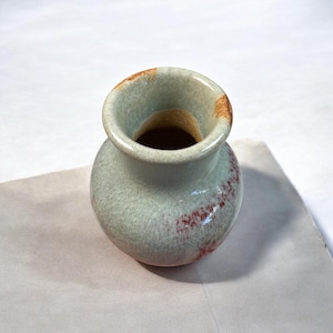 Könnte beinhalten: Kleine, runde Keramikvase mit weiter Öffnung. Die Vase hat eine hellgrüne Glasur mit rotbraunen Akzenten und einem Craquelé-Finish. Der Rand hat braune Highlights.