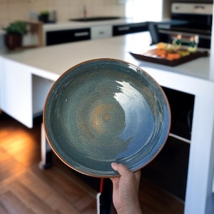 Puede incluir: Un plato de cerámica grande y redondo con un diseño de remolino azul y verde y un borde marrón. El plato se sostiene en una mano, mostrando su diseño. El plato es probablemente para servir comida.
