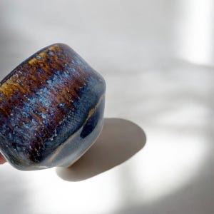 May include: A handmade ceramic bowl with a unique glaze. The bowl features a blend of blue, brown, and gold colors, creating a textured, iridescent effect. The bowl's shape is slightly asymmetrical, adding to its handcrafted appeal.