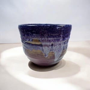 May include: A handmade ceramic bowl with a unique glaze. The bowl features a dark blue rim that fades into shades of purple, gold, and light blue. The glaze has a dripped effect, adding texture and visual interest to the piece.